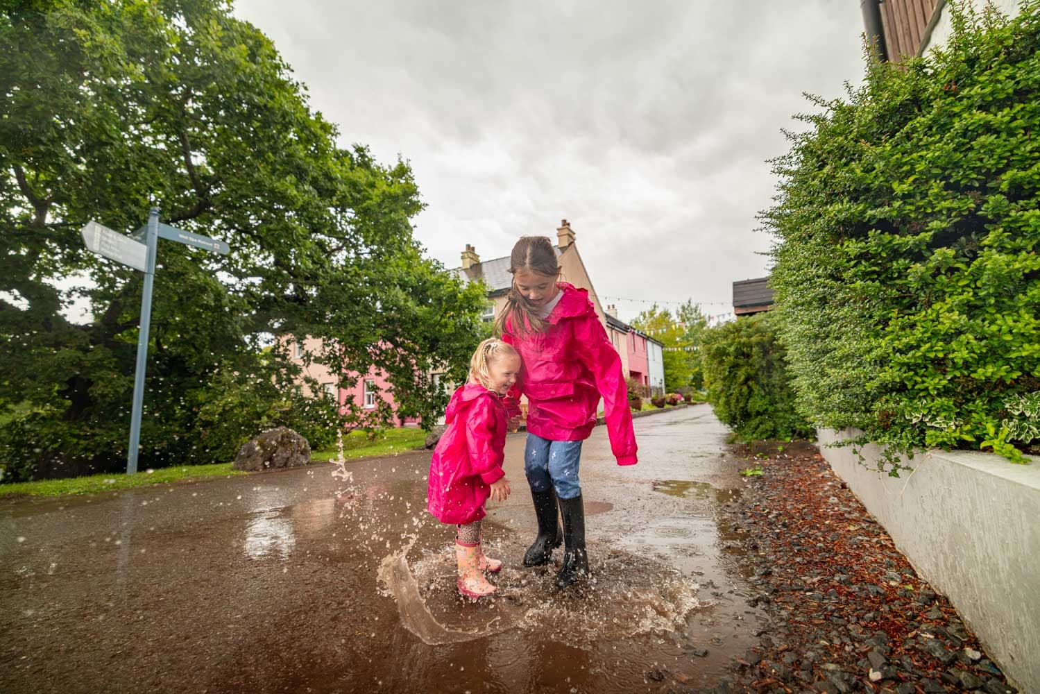 Girl And Toddler Splashing In Puddle Village Autumn